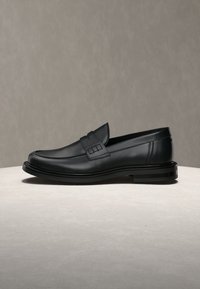 Chaussure penny loafer en cuir noir avec un petit talon, posée sur une surface claire sur un fond gris neutre.