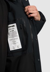 Juodos striukės vidus, turintis užtraukiamą kišenę, etiketę su priežiūros instrukcijomis ir glotnų, matinį paviršių. Matomi užtrauktuko ir spaustuko uždarymo detalės.