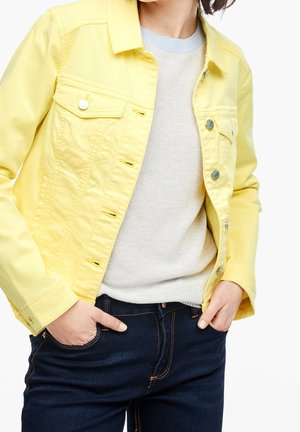 Person trägt eine hellgelbe Jeansjacke über einem hellbeigen Strickpullover mit dunkelblauen Jeans, Hände in den Taschen, Ausschnitt auf Schulter- und Taillenhöhe.