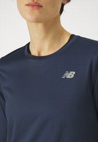 Granatowa sportowa koszulka z teksturowaną tkaniną, okrągłym dekoltem i odblaskowym srebrnym logo na lewej stronie klatki piersiowej.