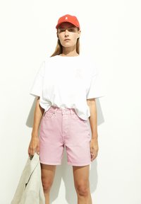 Korte mouwen witte t-shirt met een lichtroze lintlogo, gecombineerd met lichtroze denim shorts en een rode pet. Houdt een beige jas vast.