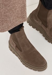 Brown suede ankle boots with elastic side panels and chunky rubber soles, featuring a textured finish and stitched details along the edges.