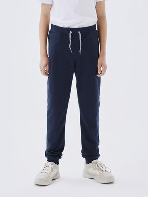 Pantalons de jogging bleu marine avec taille élastique, cordon de serrage et poches zippées, assortis à des baskets de couleur claire. Texture lisse.