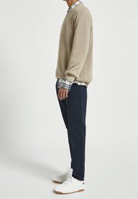 Pull en tricot côtelé beige superposé sur une chemise à carreaux bleus et blancs, associé à un pantalon noir marine et des baskets blanches.