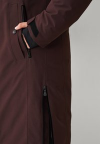 Bordeauxrode waterdichte jas met een gladde textuur, ritszakken en verstelbare manchetten, voorzien van zwarte ritsen en klittenbanddetails.