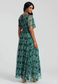Robe maxi florale verte avec des manches transparentes, une taille froncée et un détail en forme de trou de serrure au dos, en tissu léger et texturé.