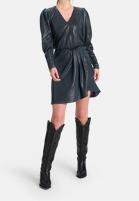 Błyszcząca turkusowa sukienka kopertowa z długimi rękawami, dekoltem w kształcie litery V i bocznym rozcięciem, zestawiona z czarnymi botkami na wysokim obcasie do kolan.