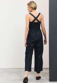 Jeans-Overall mit überkreuzten Trägern, dunkelblau, lockere Passform, verkürzte Beine und elastischem Bund. Begleitet von braunen Slippers.