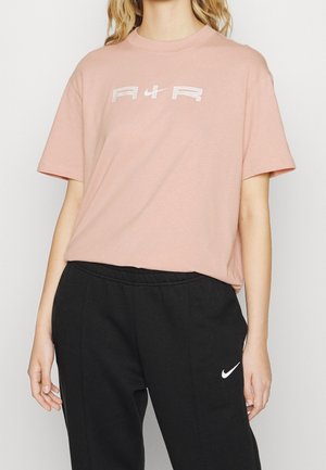 Kobieta ubrana w jasnoróżowy T-shirt z napisem "AIR" oraz czarne spodnie dresowe Nike z białym logo Swoosh, stojąca na jednolitym tle.