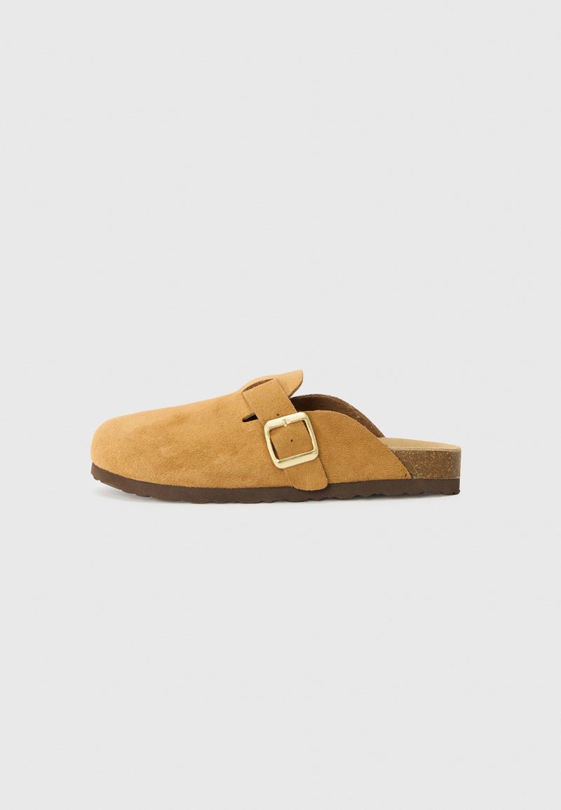 Brune ruskind klipklapper med en sidelås og detaljer i guld. Flad sål med tekstureret finish, åben bag og rundt tådesign.