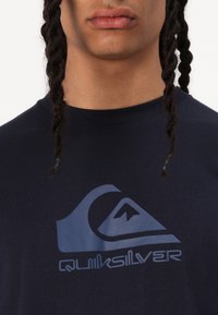 Tumši zila kokvilnas T-krekls ar Quiksilver logo gaiši zilā krāsā, apaļu kakla izgriezumu un īsām piedurknēm. Vienmērīga tekstūra, ikdienišķs dizains.