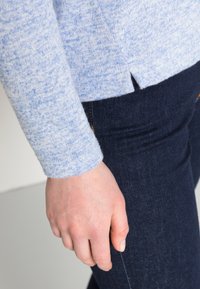 Suéter azul claro, texturizado, com mangas longas, apresentando uma ligeira fenda na bainha, combinado com jeans de couro escuro.