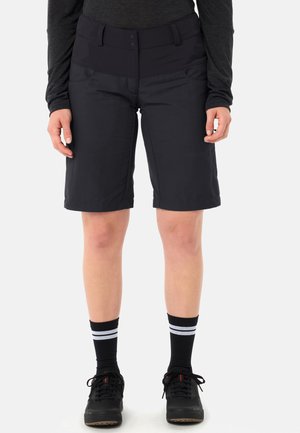 Personne portant un short noir au-dessus du genou, un haut à manches longues noir, des chaussures noires et des chaussettes noires à rayures blanches, debout sur un fond blanc.