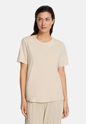 Donna con capelli scuri che indossa una T-shirt beige semplice a maniche corte e pantaloni beige traforati, in piedi su uno sfondo bianco.