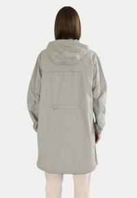 Giacca impermeabile grigio chiaro con cappuccio, maniche lunghe e taglio a lunghezza intermedia. Presenta una tasca sul retro con zip e polsini elasticizzati.