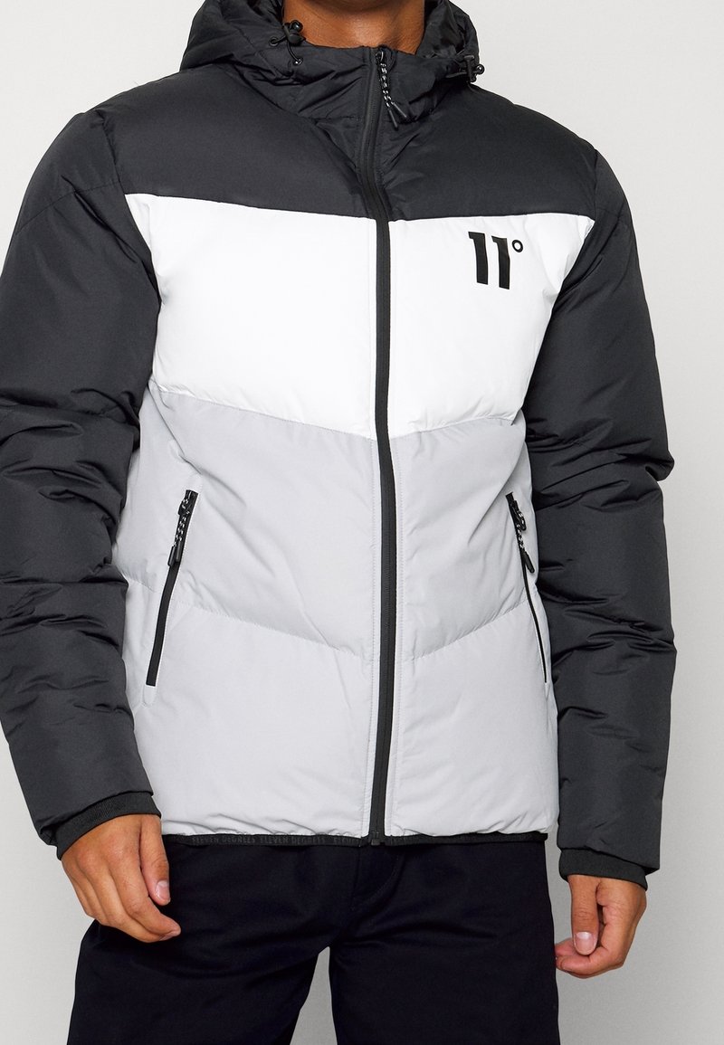 Homme portant une veste rembourrée noire, blanche et grise avec capuche et fermeture éclair à l'avant, affichant le logo "11°" sur la poitrine.