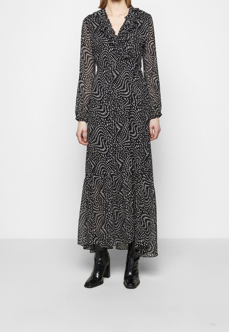 Vrouw draagt een zwarte maxi-jurk met wit abstract gestippeld patroon, lange mouwen, een V-hals met ruches en zwarte enkellaarzen met hak.
