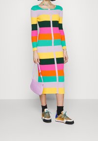 Vestido de punto con llamativas rayas horizontales en rosa, verde, amarillo, naranja y púrpura, con un cierre frontal de botones. Combinado con zapatillas y un bolso rosa.