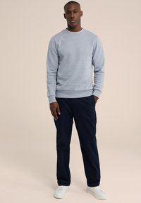 Sudadera gris claro con cuello redondo, puños y dobladillo de canalé, combinada con pantalones azul marino oscuros y zapatillas blancas, lucida por un modelo de pie.