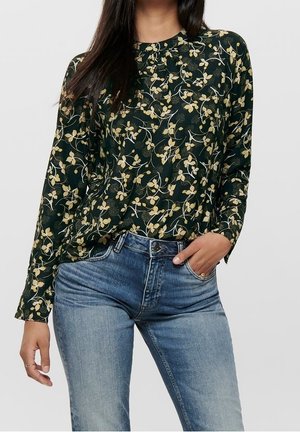 Nainen pukeutunut tummanvihreään kukalliseen pitkähihaiseen paitaan, joka on sullottu vaaleansinisiin farkkuihin, toisessa kädessä taskussa ja toinen käsi rentona.