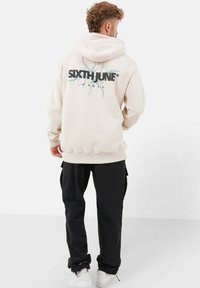 Beige Kapuzenpullover mit einem großen schwarzen und blauen Grafikdesign auf dem Rücken, auf dem der Text "SIXTH JUNE" zu sehen ist. Schwarze Cargo-Hosen und weiße Sneaker runden den Look ab.