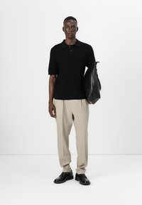 Polo noir à rayures, manches courtes, pantalon beige à plis, chaussures en cuir noires, tenant un sac noir, se tenant contre un fond blanc uni.