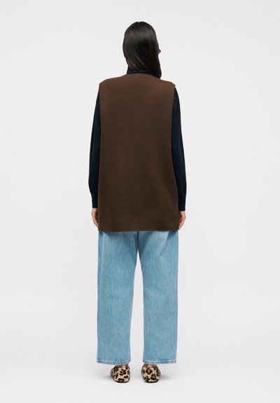 Καφέ αμάνικο πουλόβερ πάνω από σκούρο τοπ, συνδυασμένο με ανοιχτού τόνου μπλε τζιν με φαρδιά παντελόνια και παπούτσια με λεοπάρ σχέδιο. Λεπτή υφή, άνετη εφαρμογή.