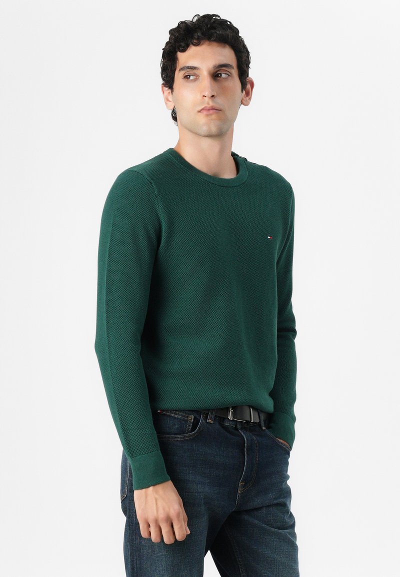 Groene gebreide trui met een textuurpatroon, ronde halslijn en lange mouwen. Voorzien van een klein logo op de borst en gecombineerd met donkere spijkerbroeken.