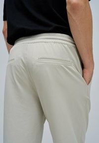 Pantalons de jogging beige clair avec une taille élastique, présentant des poches arrière et une texture de tissu lisse et légèrement brillante.