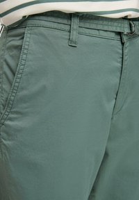 Pantalon en coton vert avec poche avant et passants de ceinture, porté avec une chemise rayée rentrée.