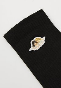 Juodas širdies rašto medis su maža balta siuvinėta stilizuota veido emblema, papuošta auksinėmis ir persikinėmis detalėmis.