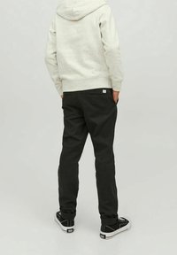 Person som bär en ljusgrå hoodie, svarta byxor och svarta sneakers, står med händerna i bakfickorna och vänder bort.