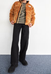 Chaqueta de cremallera marrón y esponjosa con detalles en negro; camiseta de rayas de manga larga debajo; pantalones de pierna ancha negros; botas negras.