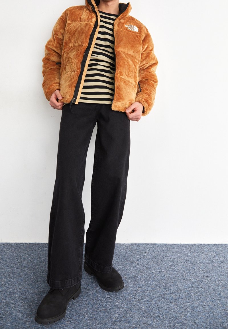 Chaqueta de cremallera marrón y esponjosa con detalles en negro; camiseta de rayas de manga larga debajo; pantalones de pierna ancha negros; botas negras.
