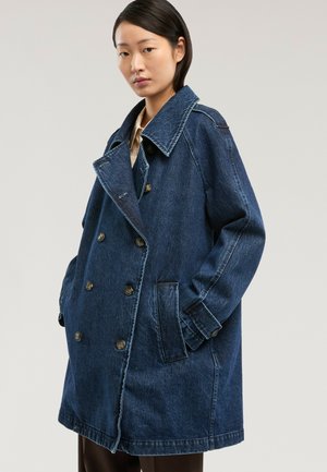 Mujer con abrigo vaquero azul oscuro de doble botonadura y cuello ancho, de gran tamaño, con las manos en los bolsillos, sobre un fondo claro y liso.