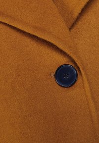 Wollen mantel in mosterdgeel, voorzien van een grote marineblauwe knoop met vier gaatjes, een reverskraag en een zacht, gestructureerd oppervlak.