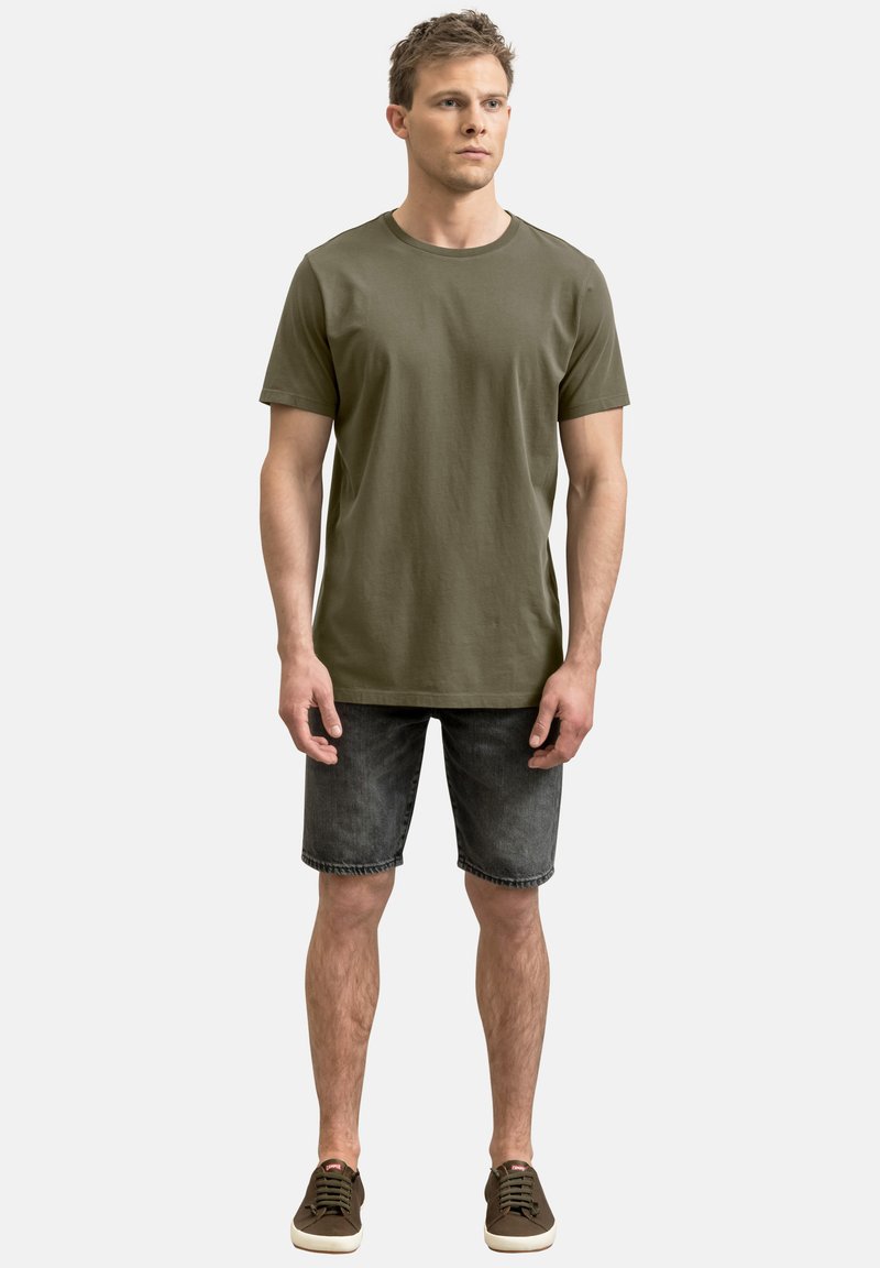 T-shirt in cotone verde oliva con maniche corte, abbinato a pantaloni corti in denim scuro. L'outfit è completato da scarpe stringate marroni.