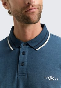 Blaues Poloshirt mit strukturiertem Stoff, einem weiß-blauen gestreiften Kragen, einer Drei-Knopf-Leiste und einem Logo mit "1962" auf der Brust.