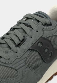 Gros plan d'une sneaker grise avec un tissu texturé, des surpiqûres en daim, des lacets gris et un patch logo noir sur le côté.