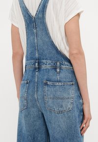 Gaišie zilie denim kombinezoni ar V formas kakla dizainu aizmugurē, regulējamas siksnas un divām aizmugurējām kabatām ar zeltījuma šuvēm.