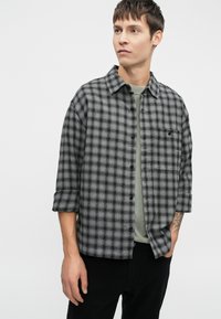 Camisa de cuadros gris y negra con un corte relajado, mangas enrolladas y un bolsillo en el pecho, llevada sobre una camiseta verde.