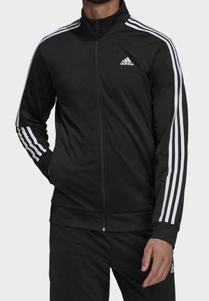 Homme portant une veste et un pantalon de survêtement Adidas noirs avec des bandes blanches et le logo Adidas sur la poitrine et la cuisse, debout devant un fond uni.