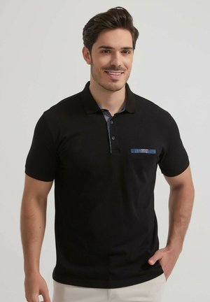 Homme souriant aux cheveux foncés portant un polo noir à manches courtes avec une bordure à motif sur la poche et le col, debout devant un fond uni.