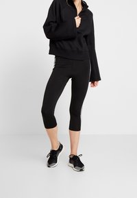 Sudadera negra recortada con diseño de media cremallera, combinada con leggins capri negros con franjas laterales blancas y zapatos deportivos negros.