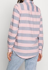 Une personne portant une chemise à manches longues avec de larges rayures horizontales rose et grises, et un jean en denim bleu clair, vue de dos.