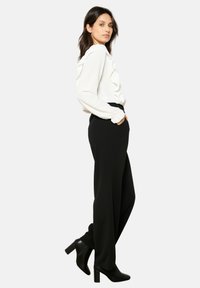 White long-sleeve blouse with ruffled detail, paired with high-waisted black trousers. Model wears black high-heeled ankle boots.