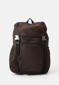 Brun nylonryggsekk med klafflokk, justerbare stropper, frontlomme med glidelås og svarte metallaksenter, med en logo på forsiden.