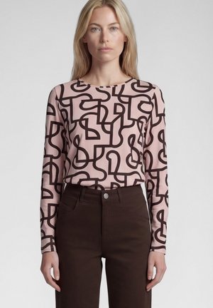 Woman wearing a long-sleeve pale pink top with abstract black line patterns and high-waisted dark brown pants standing against a plain background.