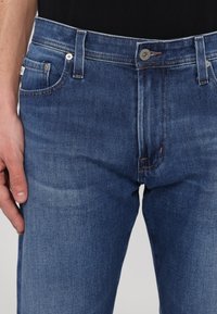 Dunkelblaue Denim-Jeans mit geradem Schnitt, leichten Abnutzungen, fünf Taschen, Kupfernähten und einem silbernen Knopfverschluss am Bund.