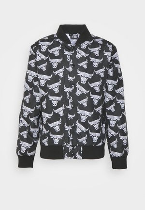 Veste bombardier noire avec motifs répétés de crânes de taureau blancs, col, poignets et taille côtelés, et fermeture à boutons-pression noirs.